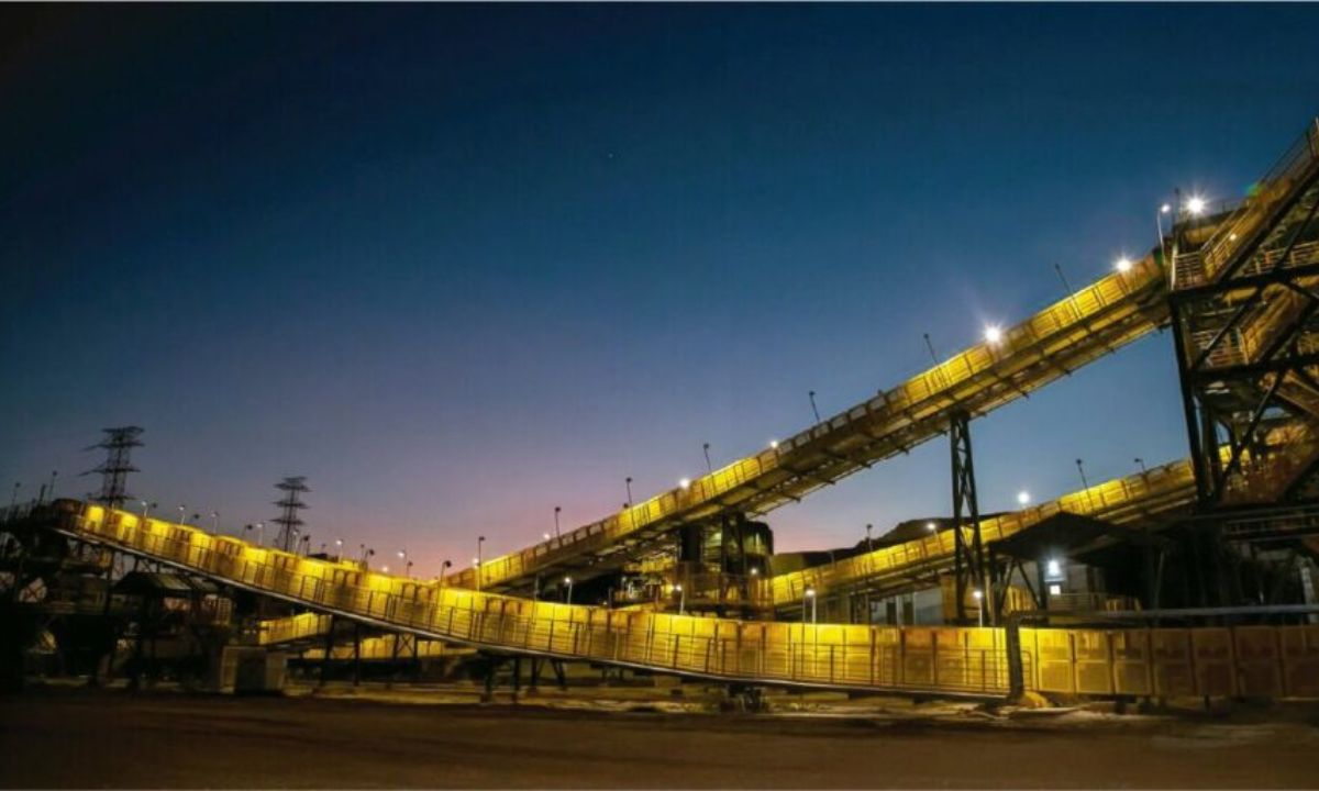 Estrutura industrial de mineração ao entardecer, com longas correias transportadoras iluminadas por luzes amarelas cruzando a imagem em diferentes alturas. O céu azul-escuro contrasta com a iluminação quente da planta, criando um cenário de grande porte e aspecto tecnológico.