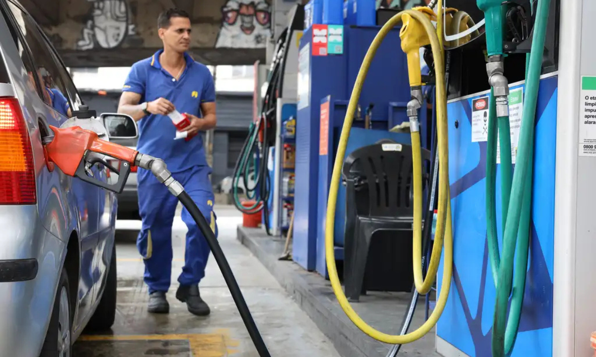Bomba de combustível ligada em carro de passeio, com frentista ao fundo.