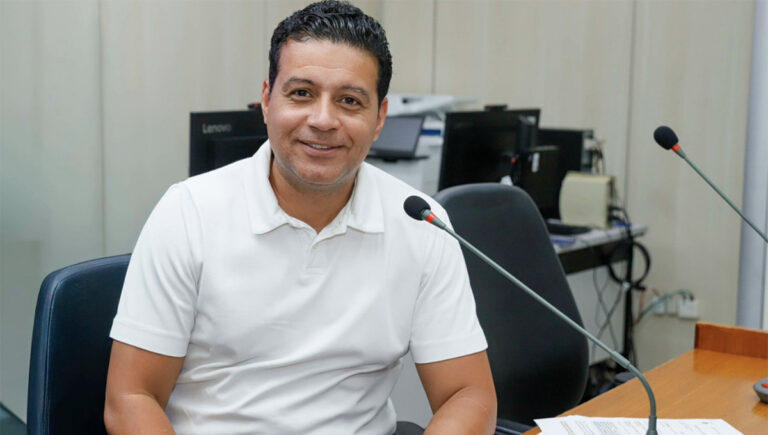 Leonardo Ângelo de camiseta branca, sorrindo, ao lado de microfone da Câmara de BH