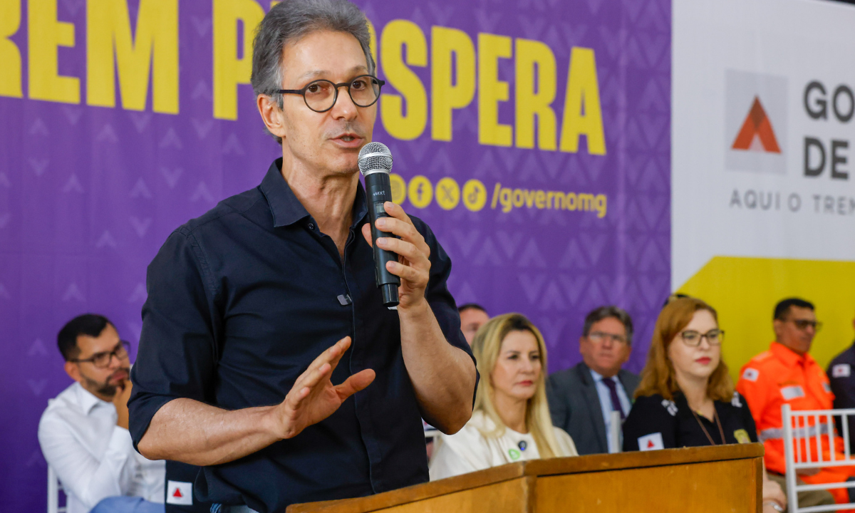 Romeu Zema com microfone na mão, à frente de backdrop do Governo de Minas