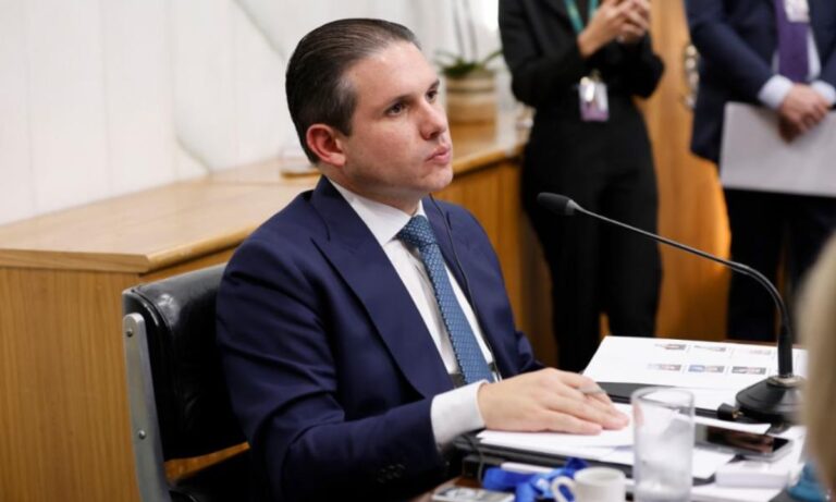 Hugo Motta, de perfil, de terno e gravata, em frente à microfone