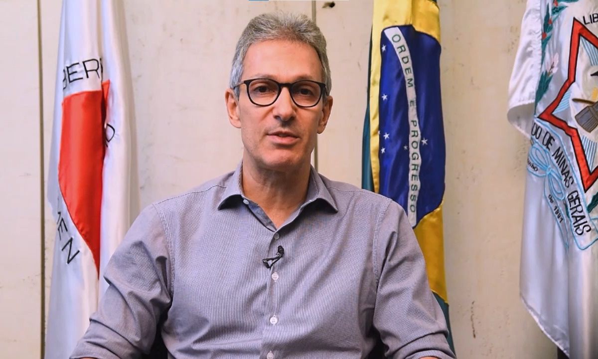 Romeu Zema de camisa social, com bandeiras de Minas Gerais e do Brasil ao fundo