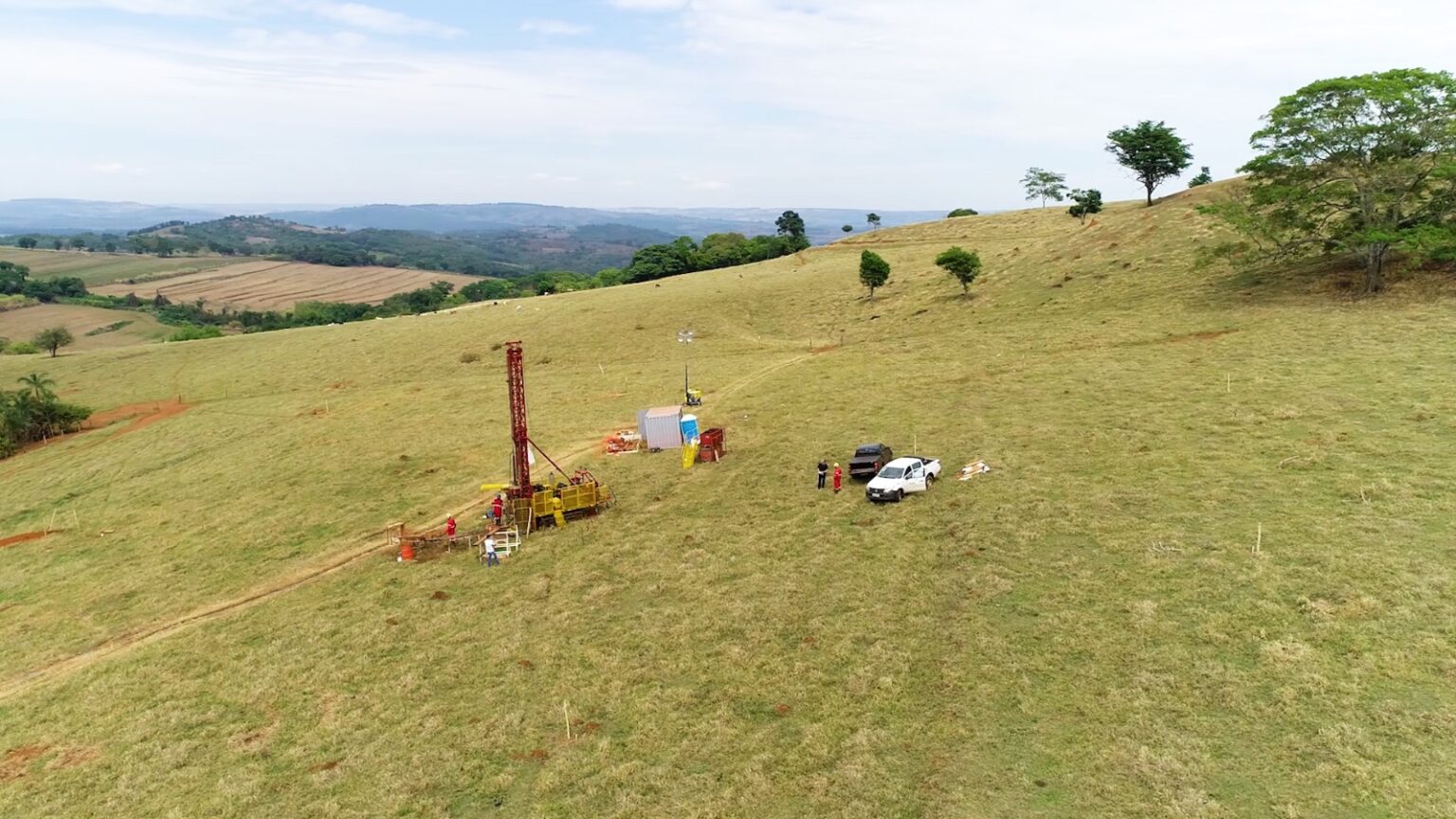 Máquinas de sondagem em meio a um campo verde rodeado de serras.