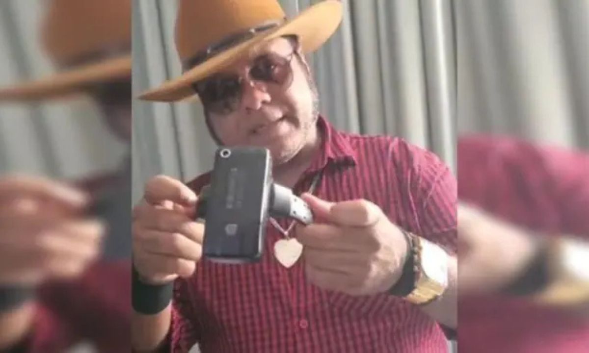 Homem de chapéu, camisa vermelha e óculos escuros segurando tornozeleira eletrônica violada