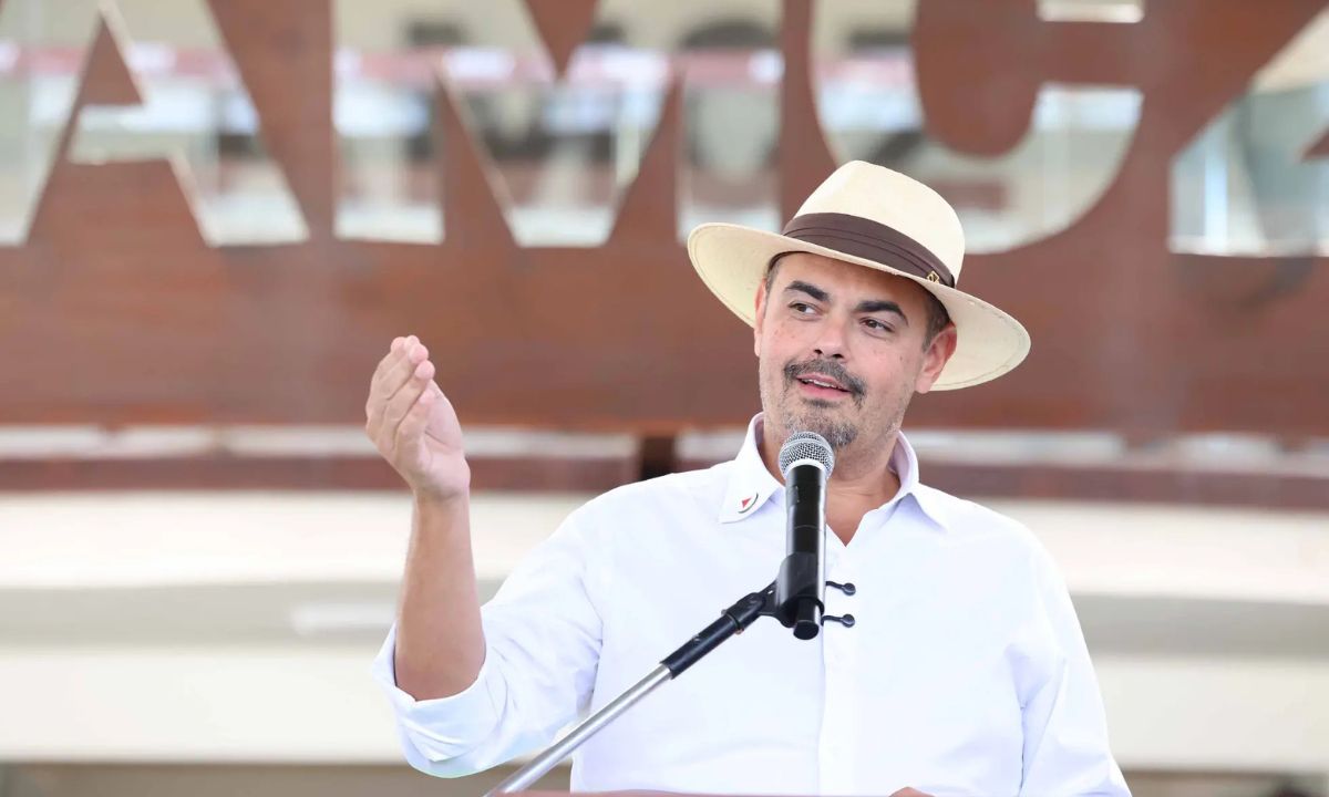 Mateus Simões falando ao microfone de chapéu, sorrindo