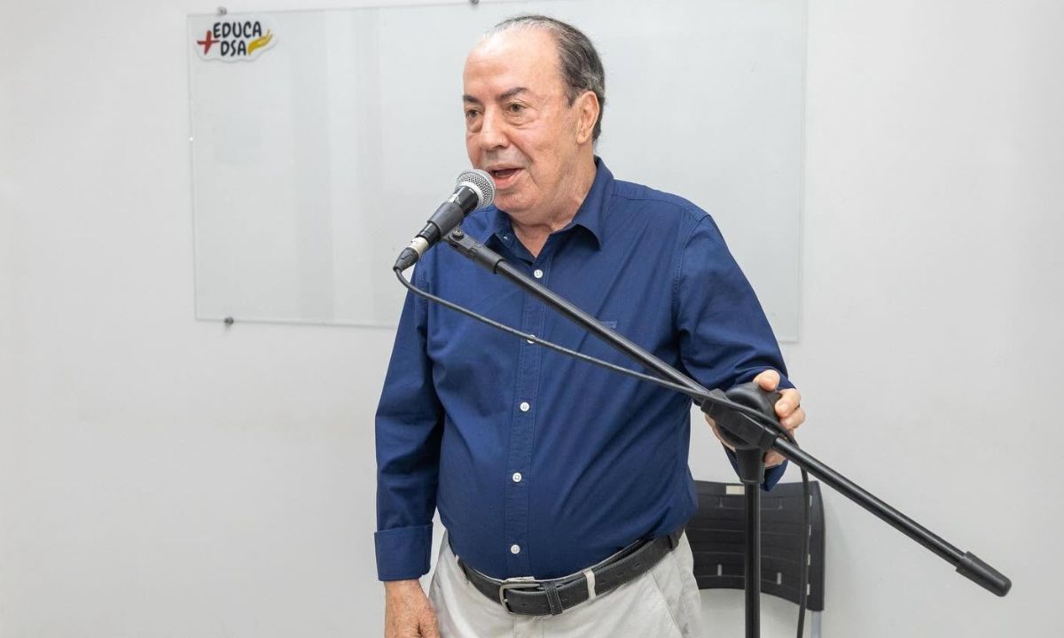 Danilo de Castro falando ao microfone de camisa social azul e calça branca