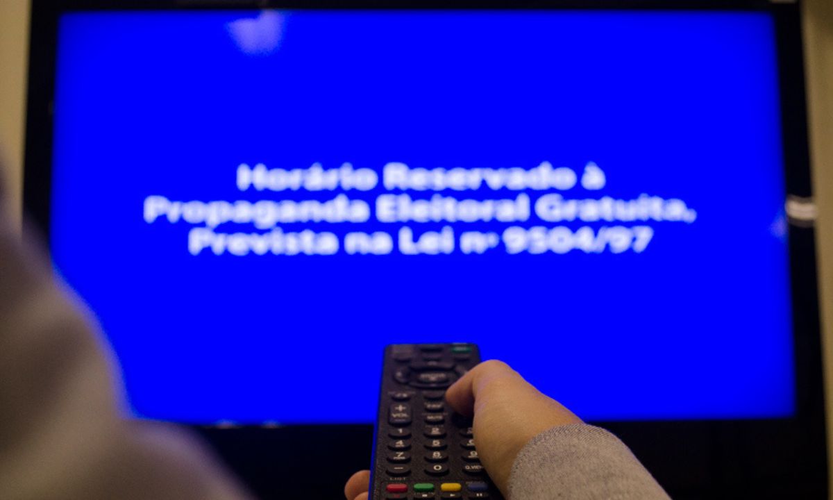 Pessoa segurando controle remoto com o aviso do horário eleitoral gratuito na TV ao fundo, desfocado