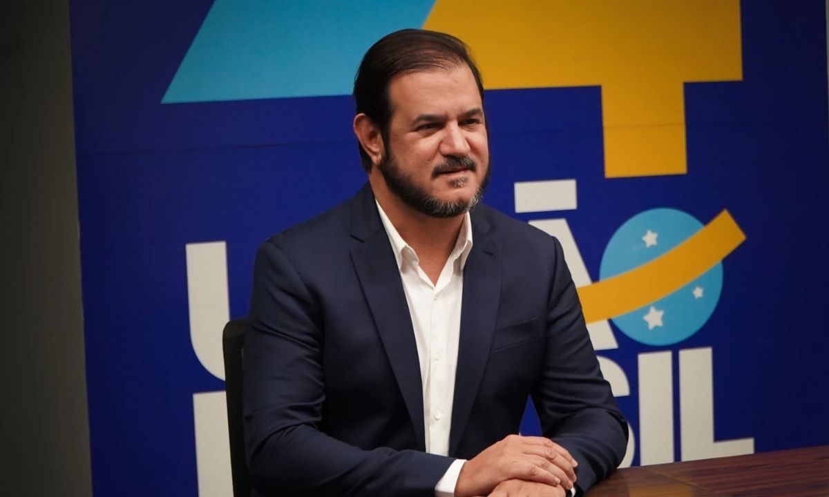 Antonio Rueda sorrindo, de terno azul, com backdrop do União Brasil ao fundo