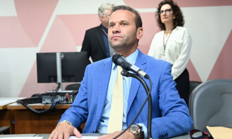 Bim da Ambulância de terno azul e gravata amarela, sentado, em frente a microfone