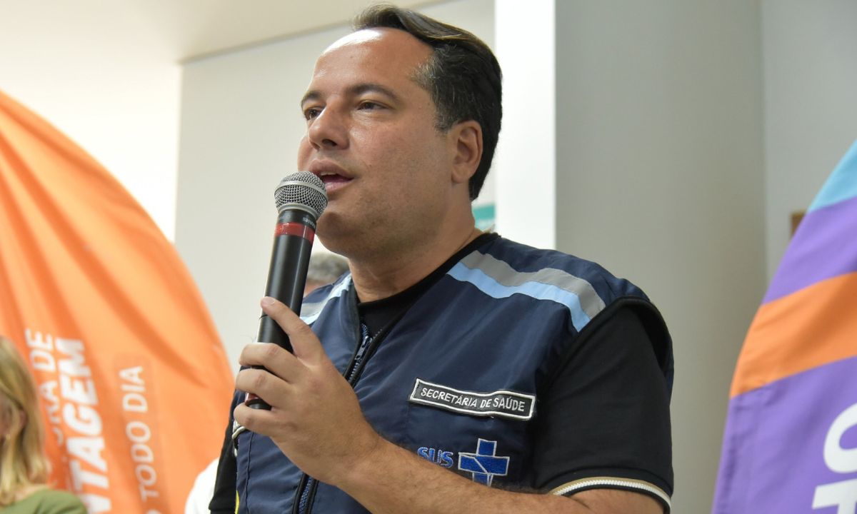 Ricardo Faria com microfone na mão esquerda, colete do SUS e camisa preta por baixo